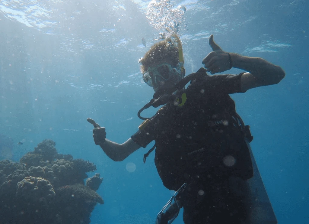 Max scuba diving underwater, giving a thumbs up over a coral reef
