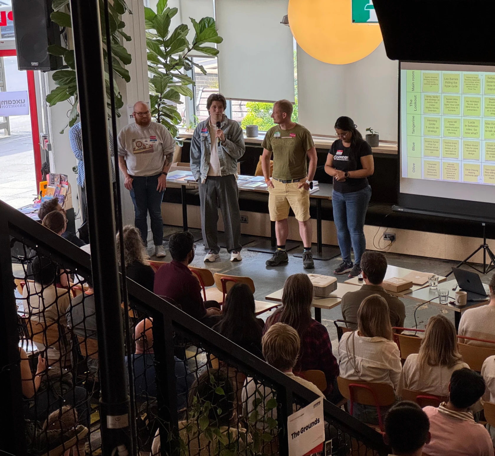 UX Camp Brighton in progress: four people stand in front of a projected session grid, one speaking into a microphone, with the audience seated on red chairs in the foreground of a plant-filled cafe space.
