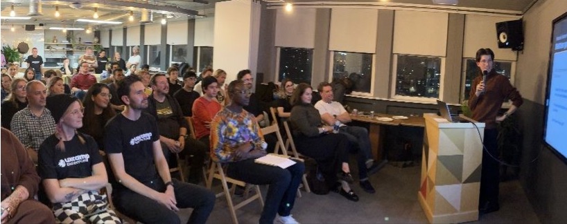 The Redux event in progress: an audience of around thirty people seated on wooden chairs in a loft office at night, watching a speaker at a wooden lectern beside a large projected slide, with the city dark through the windows behind.